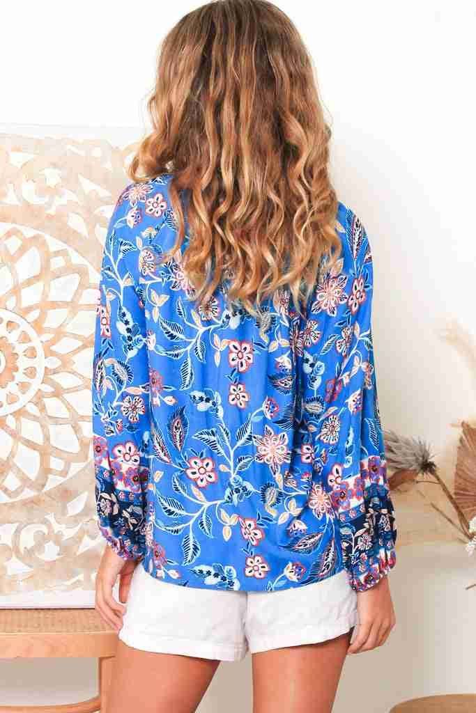 Back view of a woman wearing a blue Hana Fleur boho blouse with floral patterns and white shorts
